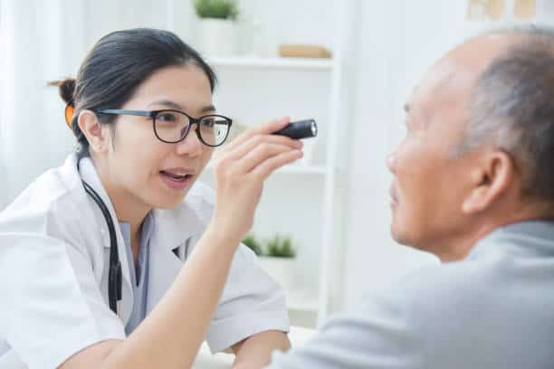 Young Asian Female Doctor wear glasses checking senior man patient eyes with flashlight in medical office.