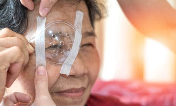 Cataract treatment after surgery by caregiver concept. Asia old woman placed protective shield over her eye taping the cover to protect during naps time.